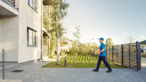 Delivery Man Holding Card Board Package Enters Through the Gates and Walks to the House. Delivering Postal Parcel. In the Background Beautiful Suburban Neighbourhood. Side View © Gorodenkoff