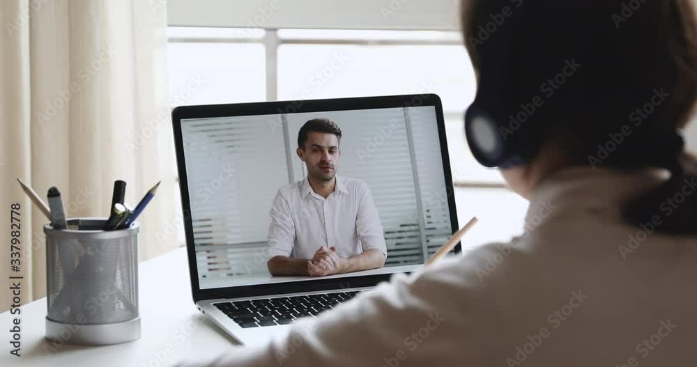 Elearning concept. Over shoulder view of student wearing headphones watching webinar with male trainer, distance learning online looking at laptop screen, writing notes. Remote education training Elearning concept. Over shoulder view of student wearing headphones watching webinar with male trainer, distance learning online looking at laptop screen, writing notes. Remote education training