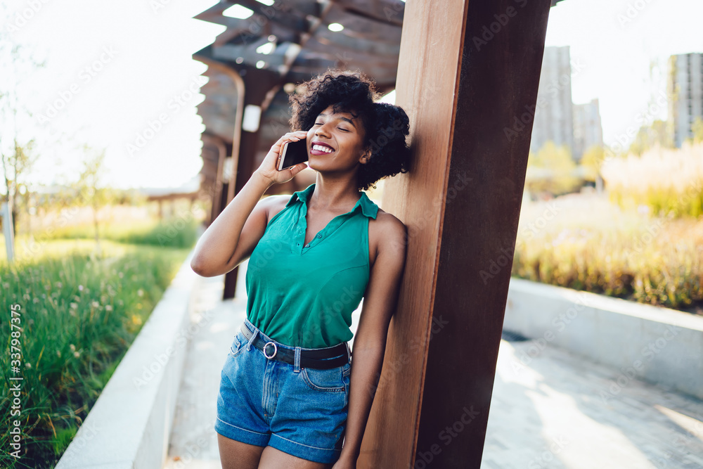Cheerful african american woman with curly hair and closed eyes having smartphone conversation standing outdoors, beautiful dark skinned female talking on mobile phone enjoy connection in roaming