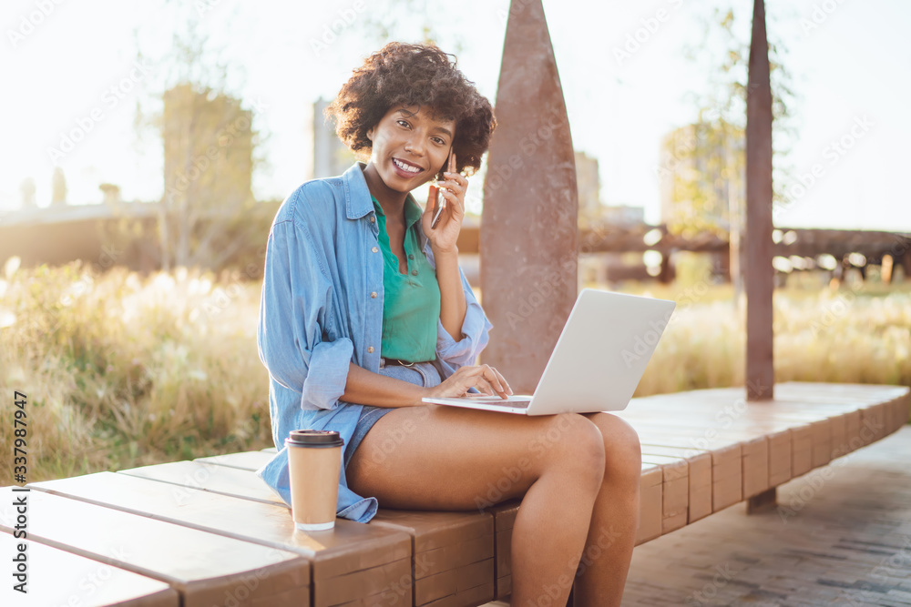 Portrait of cheerful african american woman freelancer talking on mobile phone about business online sitting in park, charming dark skinned female blogger satisfied with 4G internet use gadgets