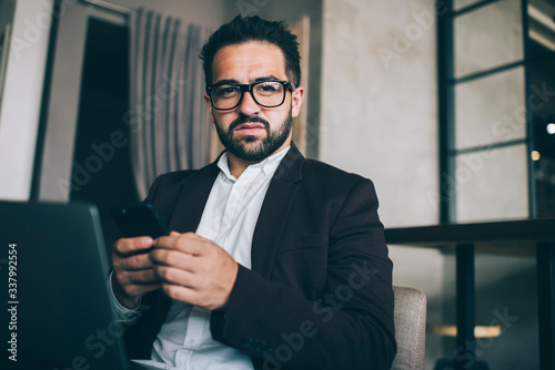 Confident serious male entrepreneur in formal outfit sending messages via app on smartphone checking exchange market news on laptop, portrait of pensive business owner look in camera with cellular