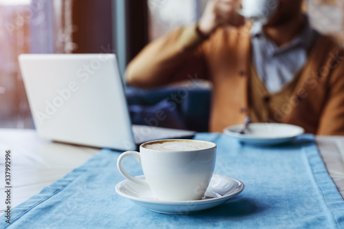 A man sits at a table and drinks coffee. A cup of coffee in the foreground. The background is blurred. A close-up.