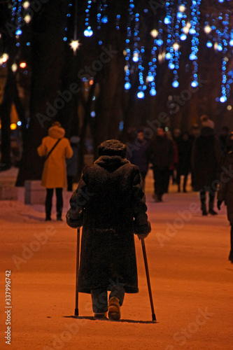 an elderly woman leaning on two sticks walks through a winter park decorated with garlands for Christmas