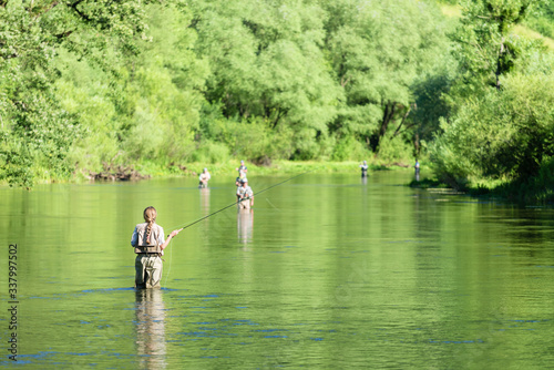 Fly fishing on the river in a rural place in the summer, young fishing woman standing in the water.Unrecognizable people in the background