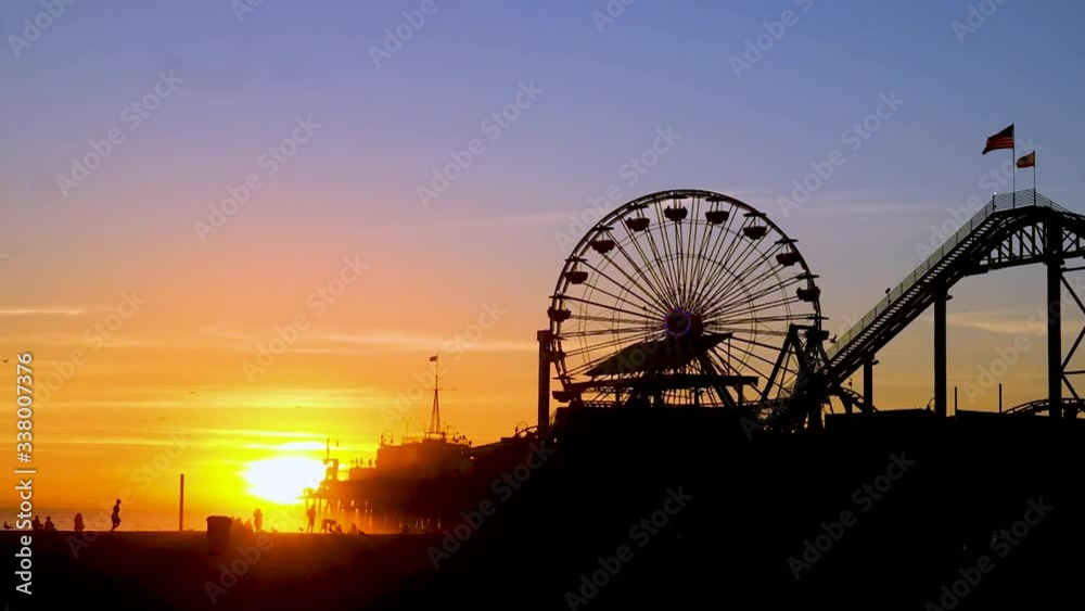 Pretty Orange Dusk Sunset Timelapse Of The Ferris Wheel And Rides On Top Of The Famous Santa Monica Pier On The Beach In California With Flags Blowing In The Wind And Seagulls Flying Around. Pretty Orange Dusk Sunset Timelapse Of The Ferris Wheel And Rides On Top Of The Famous Santa Monica Pier On The Beach In California With Flags Blowing In The Wind And Seagulls Flying Around.