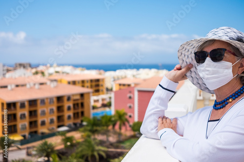 Elegantly dressed senior woman stays at home to avoid coronavirus infection, looking at the city from the balcony. Prevention and waiting for the end of the pandemic © luciano