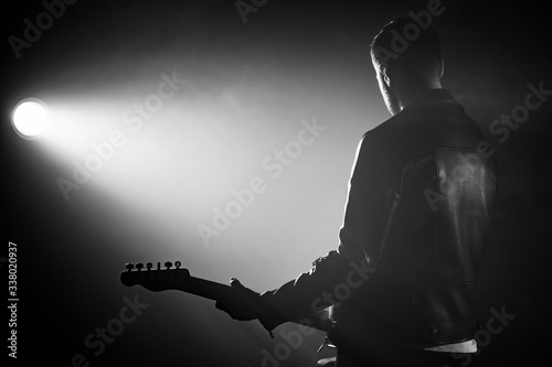 Rock guitarist man in leather jacket standing his back in smoky studio or stage masterfully playing electric guitar. View of unrecognizable musician in the spotlight. photo