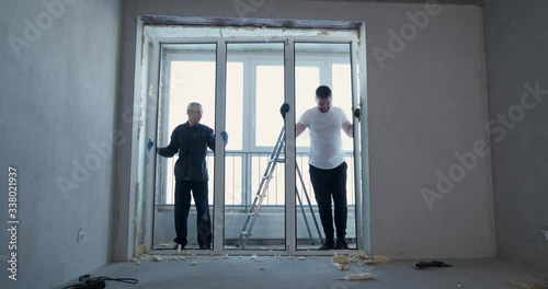 Two men at real estate object under construction dismantle plastic window, cut foam with knives, repair new apartment, replace window block, dusty work, builders, opposite window, construction site