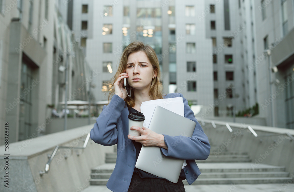 Fototapeta premium Puzzled businesswoman in front of office building having phone conversation holding laptop and documents.Female office worker with puzzled expression dealing with deadlines holding papers and computer