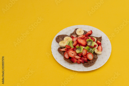 Fresh pancakes with chopped fruits, strawberries, bananas, kiwi and with honey on a light plate. Healthy food, gluten free, no eggs. Copy space. Yellow summer background. Flat lay. Top view.