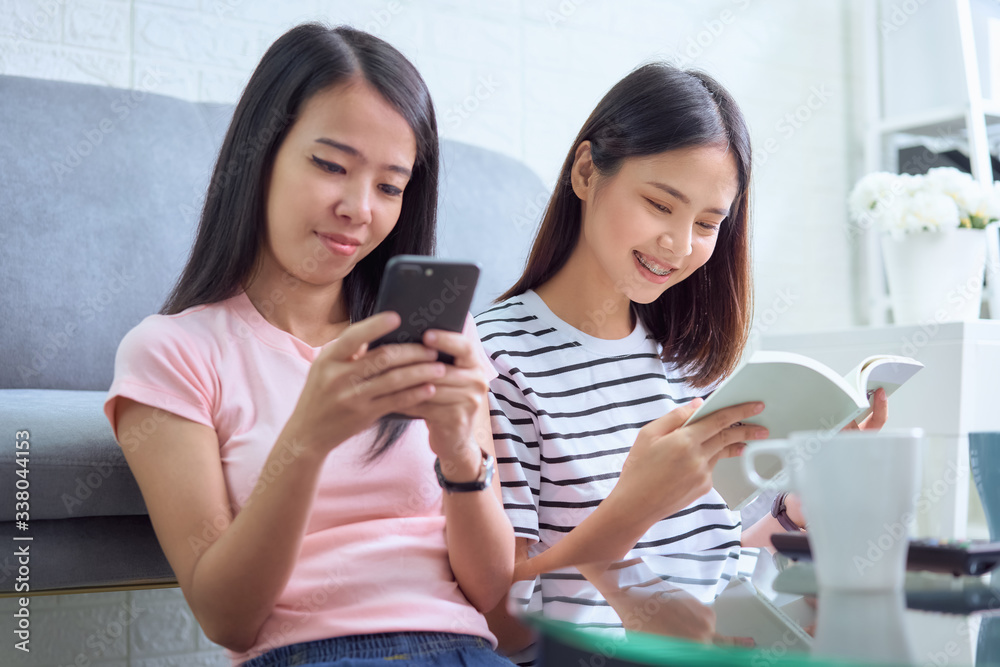 Happy young Asian woman with friends sitting on sofa and reading book, using smartphone in her living room, vacation time. Happy young Asian woman with friends sitting on sofa and reading book, using smartphone in her living room, vacation time.