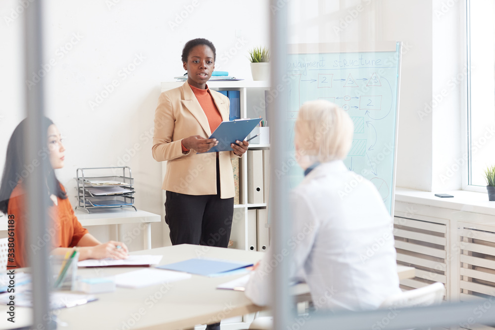 Portrait of successful African-American businesswoman giving presentation to colleagues while standing by whiteboard during meeting in conference room, copy space