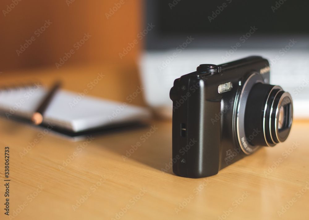 Close-up of a compact photo camera, with a laptop and notebook on a blurred background. Photography