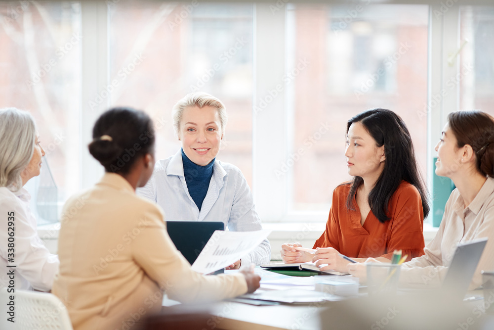 Portrait of diverse group of businesswomen discussing project while sitting at table during meeting in office, focus on smiling female boss Portrait of diverse group of businesswomen discussing project while sitting at table during meeting in office, focus on smiling female boss