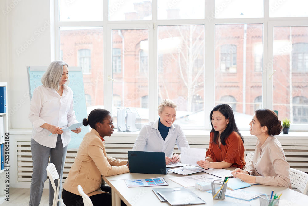 Wide angle view at diverse group of businesswomen discussing project while sitting at table against window during meeting in conference room, copy space