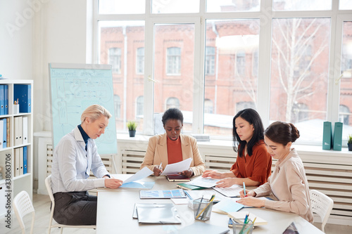 Diverse group of successful businesswomen discussing project while sitting at table against window during meeting in conference room, copy space
