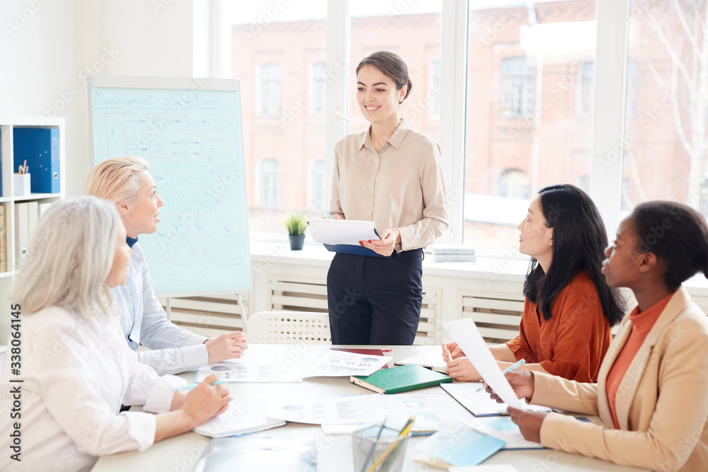 Portrait of smiling female manager presenting project plan to group of colleagues while standing by whiteboard during meeting in conference room, copy space Portrait of smiling female manager presenting project plan to group of colleagues while standing by whiteboard during meeting in conference room, copy space
