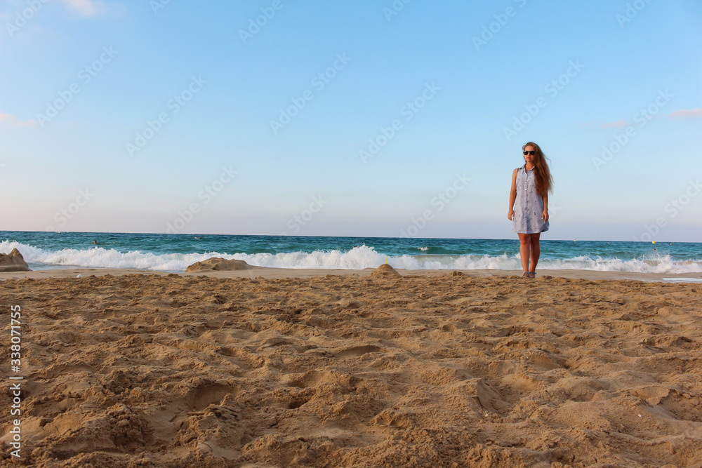 a beautiful young tourist girl with long brown unpainted hair and glasses stands tall on the beach against the background of the blue sea and Golden clean sand