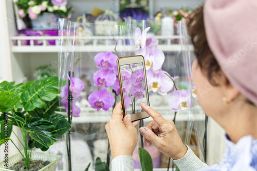 Happy caucasian elderly woman in a beret, smiling, photographs orchid flowers on the phone