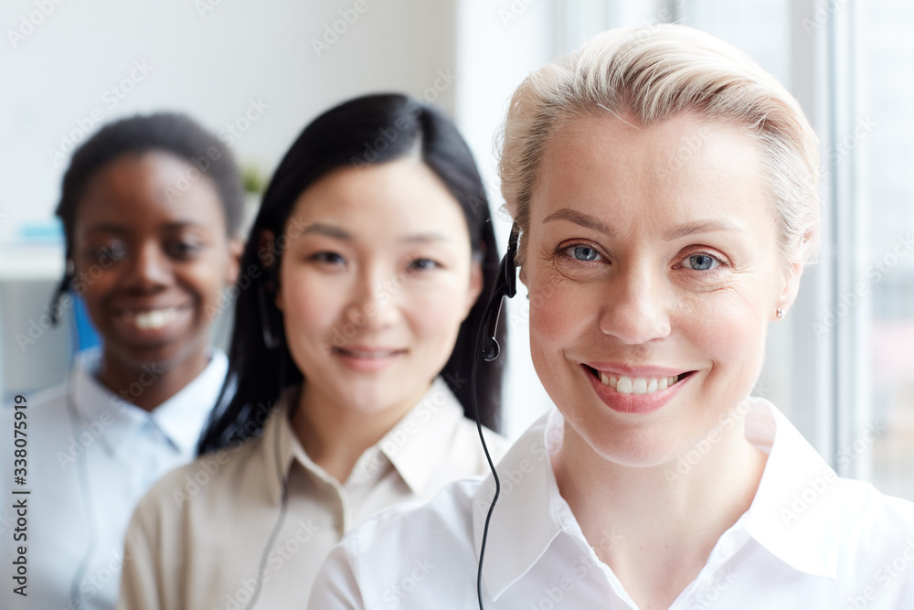 Multi-ethnic group of female call center operators looking at camera standing in row, focus on smiling blonde woman wearing headset in foreground, copy space Multi-ethnic group of female call center operators looking at camera standing in row, focus on smiling blonde woman wearing headset in foreground, copy space
