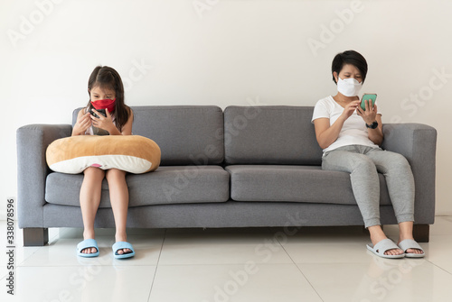 An asian mother and daughter both wearing face mask and using smert phone are sitting far from each other as social distancing concept during covid-19 or coronavirus outbreak photo