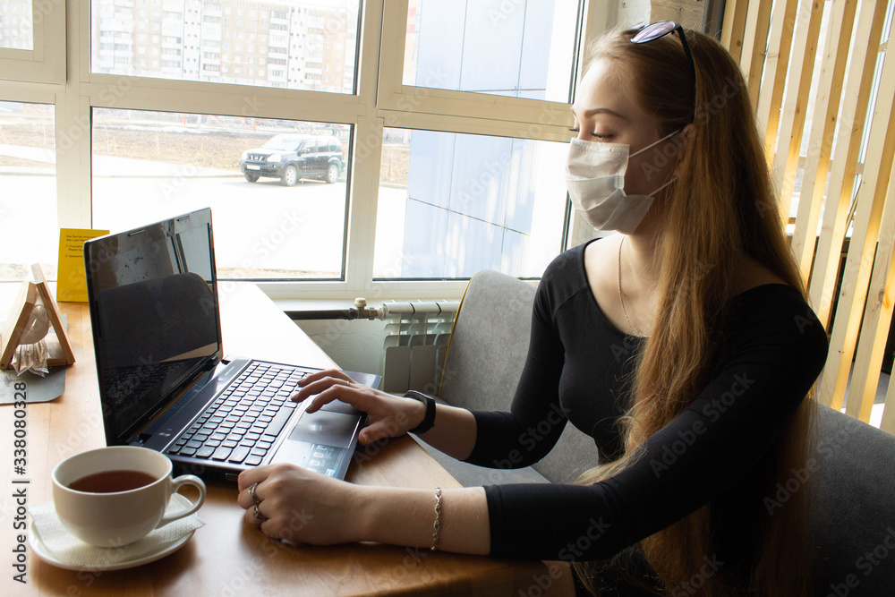Beautiful girl sits and works at the table in a medical mask. On the table computer and tea . Beautiful girl sits and works at the table in a medical mask. On the table computer and tea .