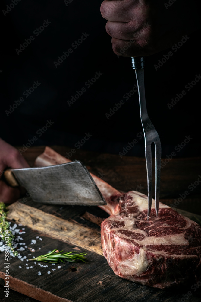 A butcher cuts beef Tomahawk steak on a bone with a cleaver on a wooden cutting Board on a dark background A butcher cuts beef Tomahawk steak on a bone with a cleaver on a wooden cutting Board on a dark background