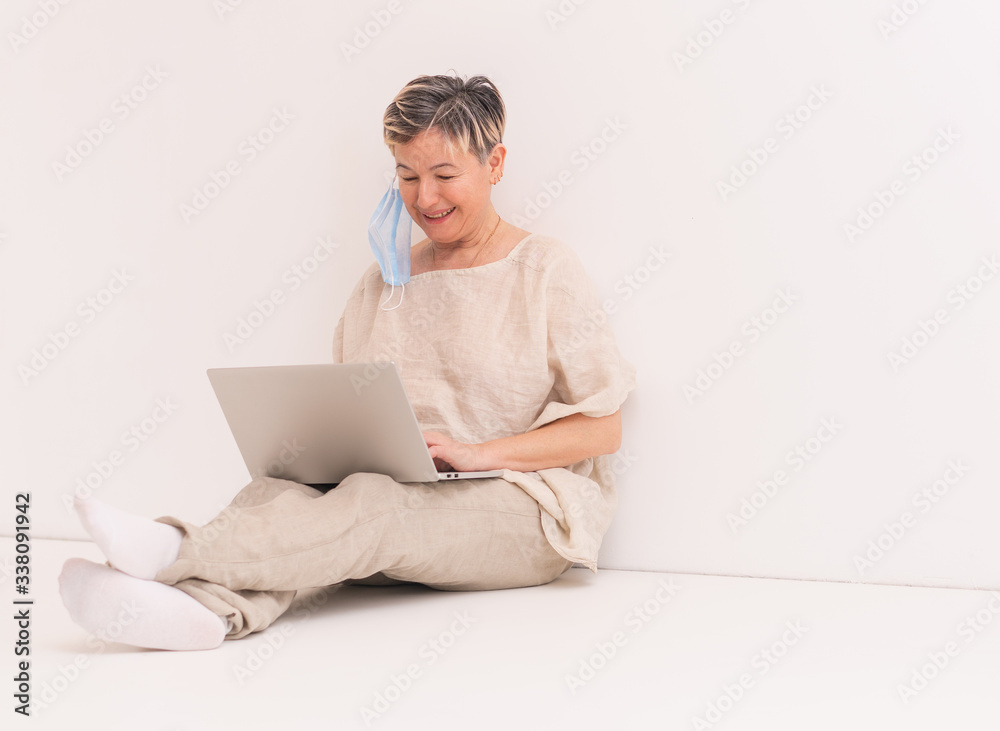 Senior happy woman with medical mask sits on the floor and working at laptop. The concept of working from home during lockdown. Copy space. Senior happy woman with medical mask sits on the floor and working at laptop. The concept of working from home during lockdown. Copy space.