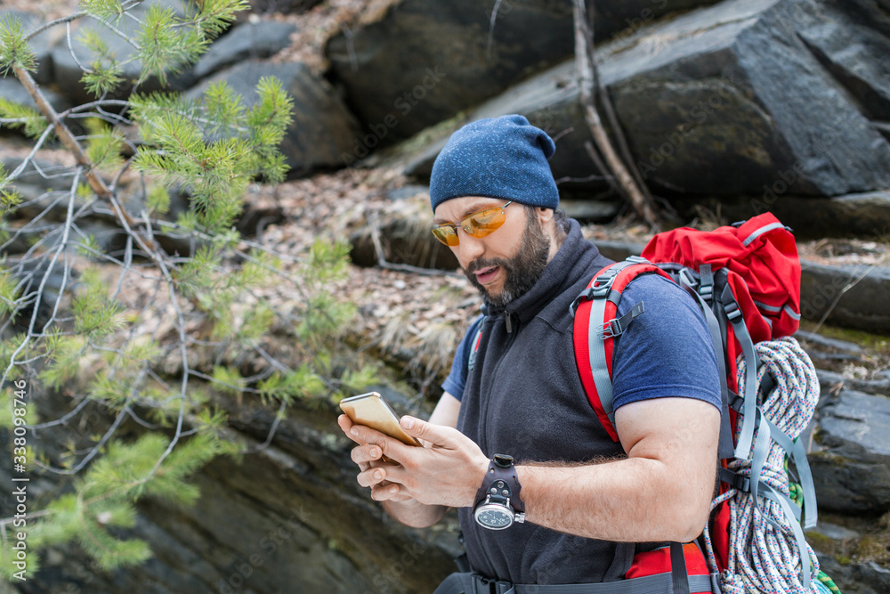 A man tourist with a backpack stands near a sheer stone rock and myotrites in the screen of a mobile phone. Searches for a way using GPS navigator.