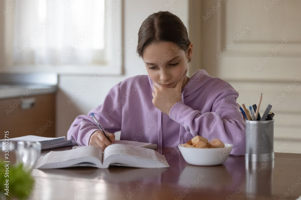 Serious teen girl school pupil studying alone sitting at kitchen table. Focused teenage student reading book, making notes doing homework at home. Adolescent tween distance learning education concept Serious teen girl school pupil studying alone sitting at kitchen table. Focused teenage student reading book, making notes doing homework at home. Adolescent tween distance learning education concept
