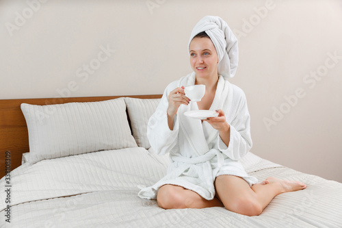 Beautiful smiling girl in white robe sitting on the bed with cup in hands