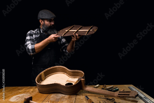 Luthier wearing cap, plaid shirt and apron. Measuring back of Spanish guitar. On the workbench guitar in construction and tools. Dark black background. photo