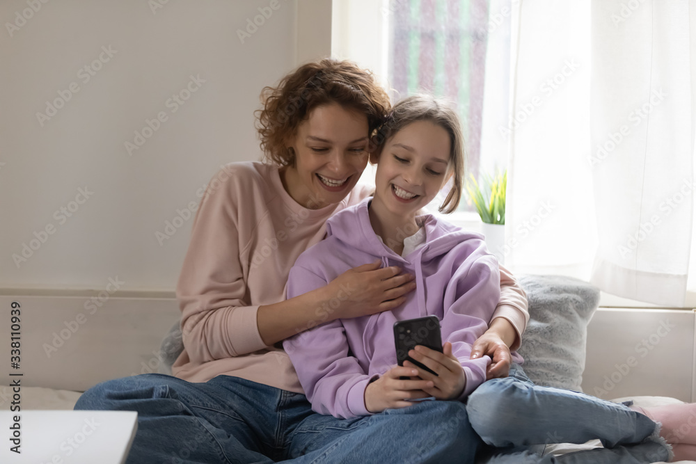 Happy teen daughter and young mum using smartphone at home taking selfie, watching funny social media video bonding at home. Smiling parent mother hugs teenager child having fun playing mobile game.