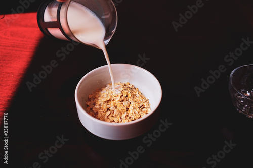 Pouring the milk into oatmeal in a white porcelain bowl on the wooden background.