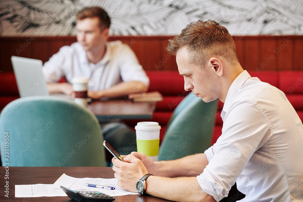 Serious frowning young businessman sitting at cafe table with documents in front of him and reading news on smartphone screen Serious frowning young businessman sitting at cafe table with documents in front of him and reading news on smartphone screen