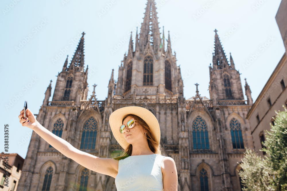 Fototapeta premium Young and happy tourist woman making selfie photo in front of cathedral in Barcelona