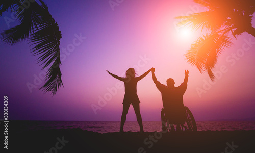 Disabled man in a wheelchair with his wife on the beach. Silhouettes at sunset