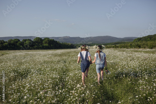 two cute teen girls in denim overalls walk in a daisy field