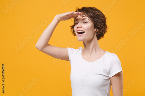 Young brunette woman girl in white t-shirt posing isolated on yellow orange wall background in studio. People lifestyle concept. Mock up copy space. Holding hand at forehead looking far away distance.