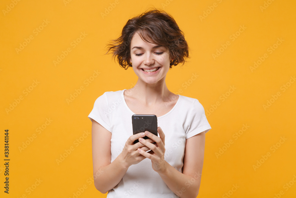 Pretty young brunette woman girl in white t-shirt posing isolated on yellow orange background studio portrait. People lifestyle concept. Mock up copy space. Using mobile phone, typing sms message. Pretty young brunette woman girl in white t-shirt posing isolated on yellow orange background studio portrait. People lifestyle concept. Mock up copy space. Using mobile phone, typing sms message.