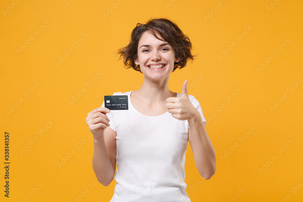 Smiling young brunette woman girl in white t-shirt posing isolated on yellow orange background studio portrait. People lifestyle concept. Mock up copy space. Hold credit bank card, showing thumb up. Smiling young brunette woman girl in white t-shirt posing isolated on yellow orange background studio portrait. People lifestyle concept. Mock up copy space. Hold credit bank card, showing thumb up.