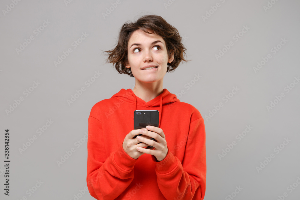 Pensive young brunette woman girl in casual red hoodie posing isolated on grey wall background studio portrait. People lifestyle concept. Mock up copy space. Using mobile phone, typing sms message. Pensive young brunette woman girl in casual red hoodie posing isolated on grey wall background studio portrait. People lifestyle concept. Mock up copy space. Using mobile phone, typing sms message.