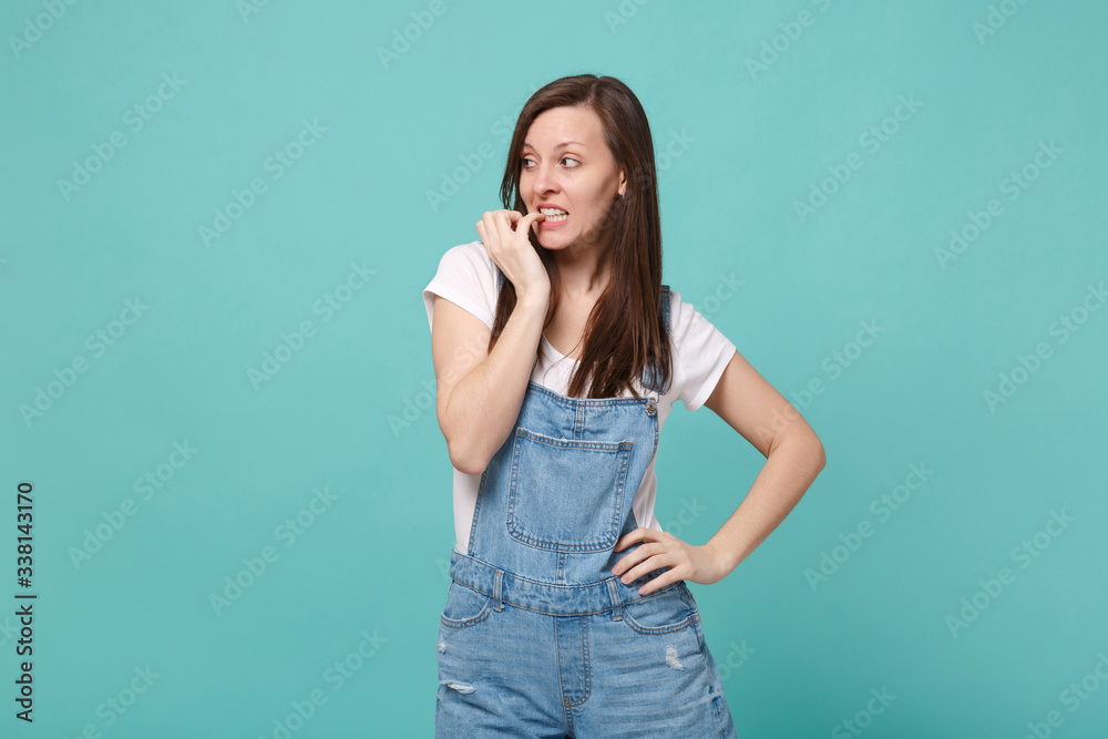 Pensive shocked young brunette woman girl in casual denim clothes posing isolated on blue turquoise background in studio. People lifestyle concept. Mock up copy space. Gnawing nails, looking aside.