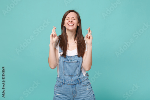 Smiling woman in casual denim clothes isolated on blue turquoise background. People lifestyle concept. Mock up copy space. Wait for special moment  keeping fingers crossed  eyes closed  making wish.
