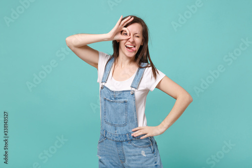 Funny young woman in casual denim clothes isolated on blue turquoise background. People lifestyle concept. Mock up copy space. Holding hands near eyes, imitating glasses or binoculars, showing tongue.