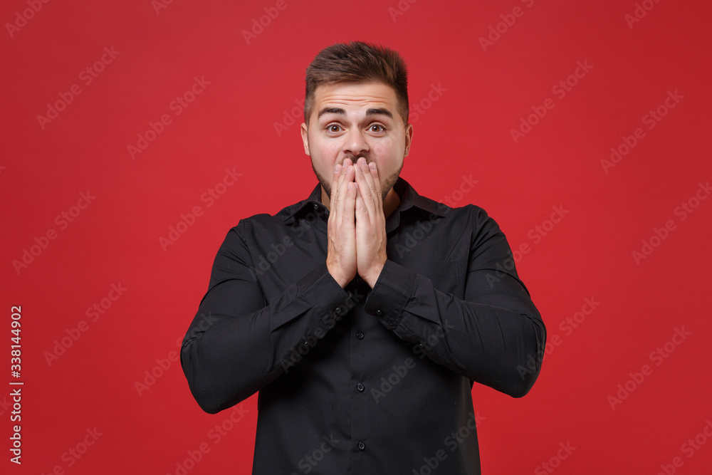 Shocked young bearded guy 20s in classic black shirt posing isolated on red wall background studio portrait. People sincere emotions lifestyle concept. Mock up copy space. Covering mouth with hands. Shocked young bearded guy 20s in classic black shirt posing isolated on red wall background studio portrait. People sincere emotions lifestyle concept. Mock up copy space. Covering mouth with hands.