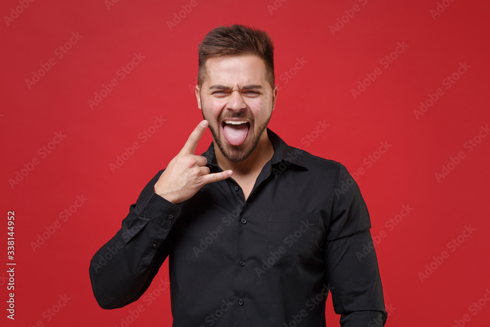 Funny young bearded guy 20s in classic black shirt posing isolated on red background. People lifestyle concept. Mock up copy space. Depicting heavy metal rock sign, horns up gesture, showing tongue. Funny young bearded guy 20s in classic black shirt posing isolated on red background. People lifestyle concept. Mock up copy space. Depicting heavy metal rock sign, horns up gesture, showing tongue.