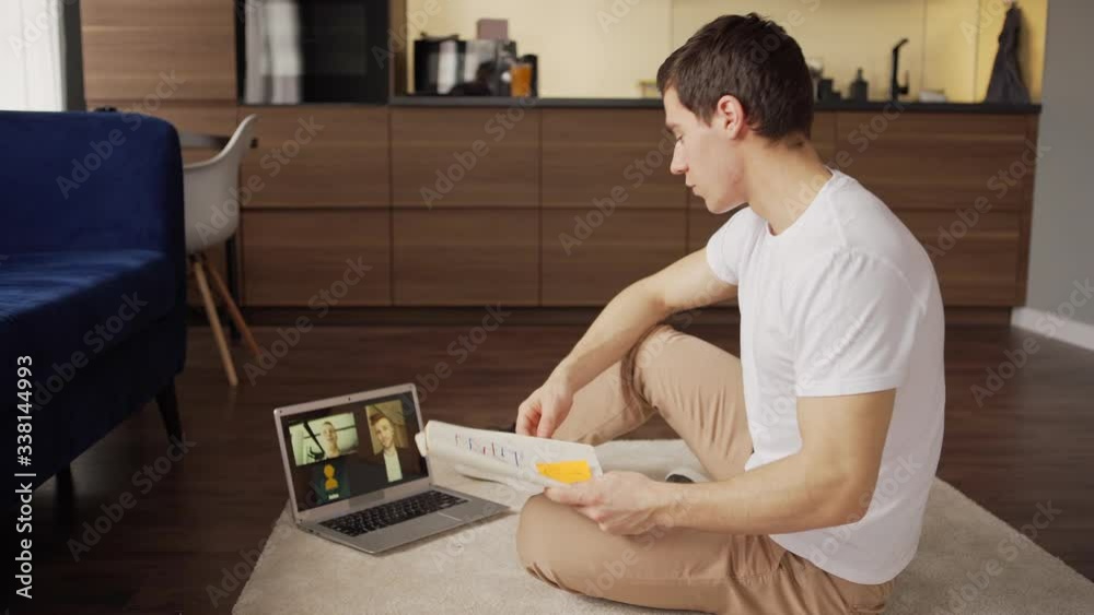 Tilt up side view of young man discussing ideas with two business partners during video conference on laptop computer sitting on floor at home Tilt up side view of young man discussing ideas with two business partners during video conference on laptop computer sitting on floor at home