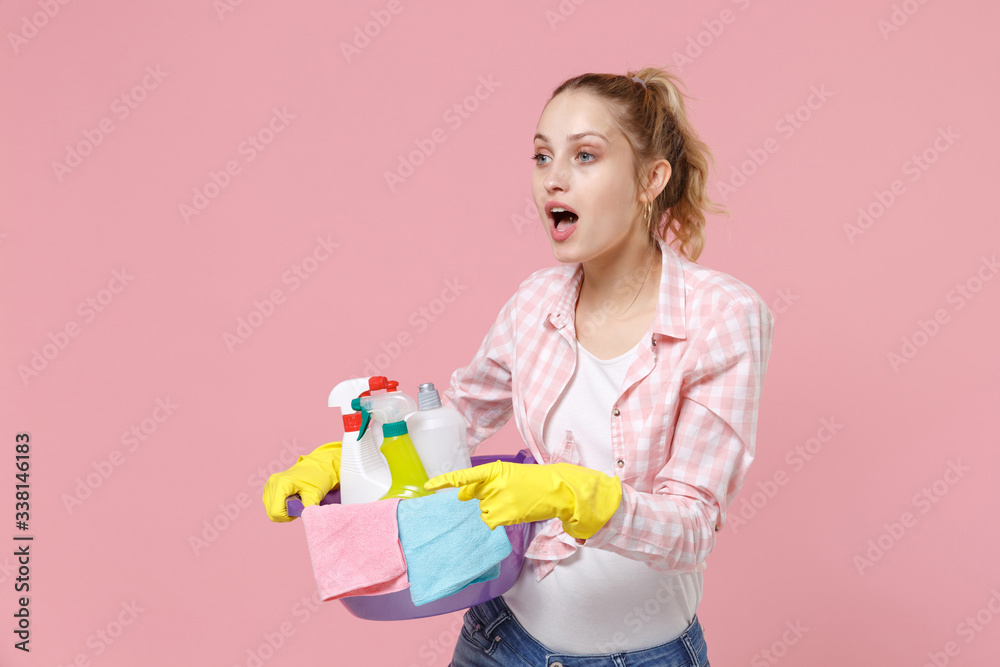 Shocked young woman housewife in rubber gloves hold basin with detergent bottles washing cleansers doing housework isolated on pink background studio. Housekeeping concept. Point index finger aside. Shocked young woman housewife in rubber gloves hold basin with detergent bottles washing cleansers doing housework isolated on pink background studio. Housekeeping concept. Point index finger aside.