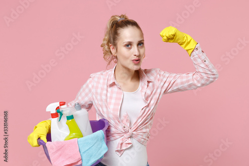 Strong young woman housewife in rubber gloves hold basin with detergent bottles washing cleansers doing housework isolated on pink background studio. Housekeeping concept. Showing biceps, muscles.